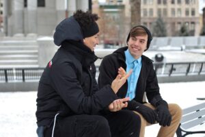 Two young men smiling and talking to each other on a bench in the snow